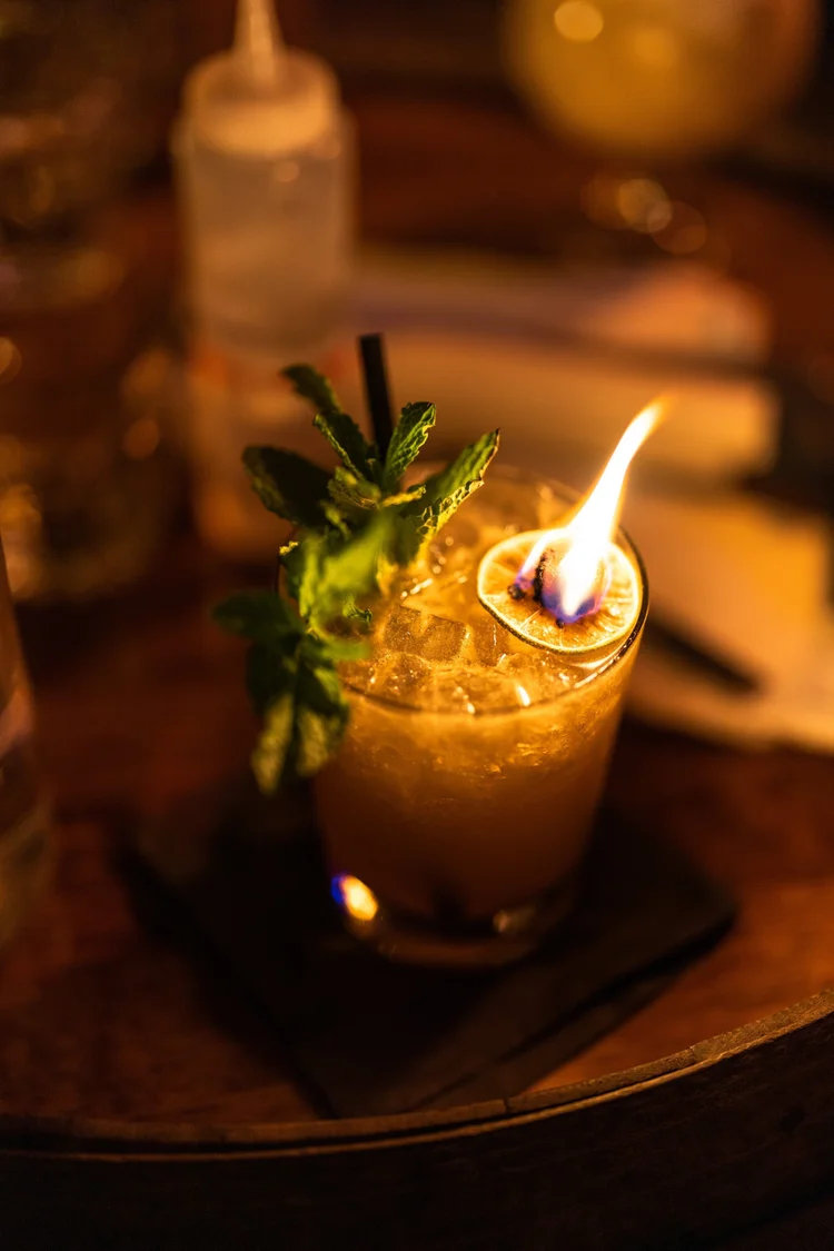 A warmly lit cocktail with a flaming garnish on a dark bar