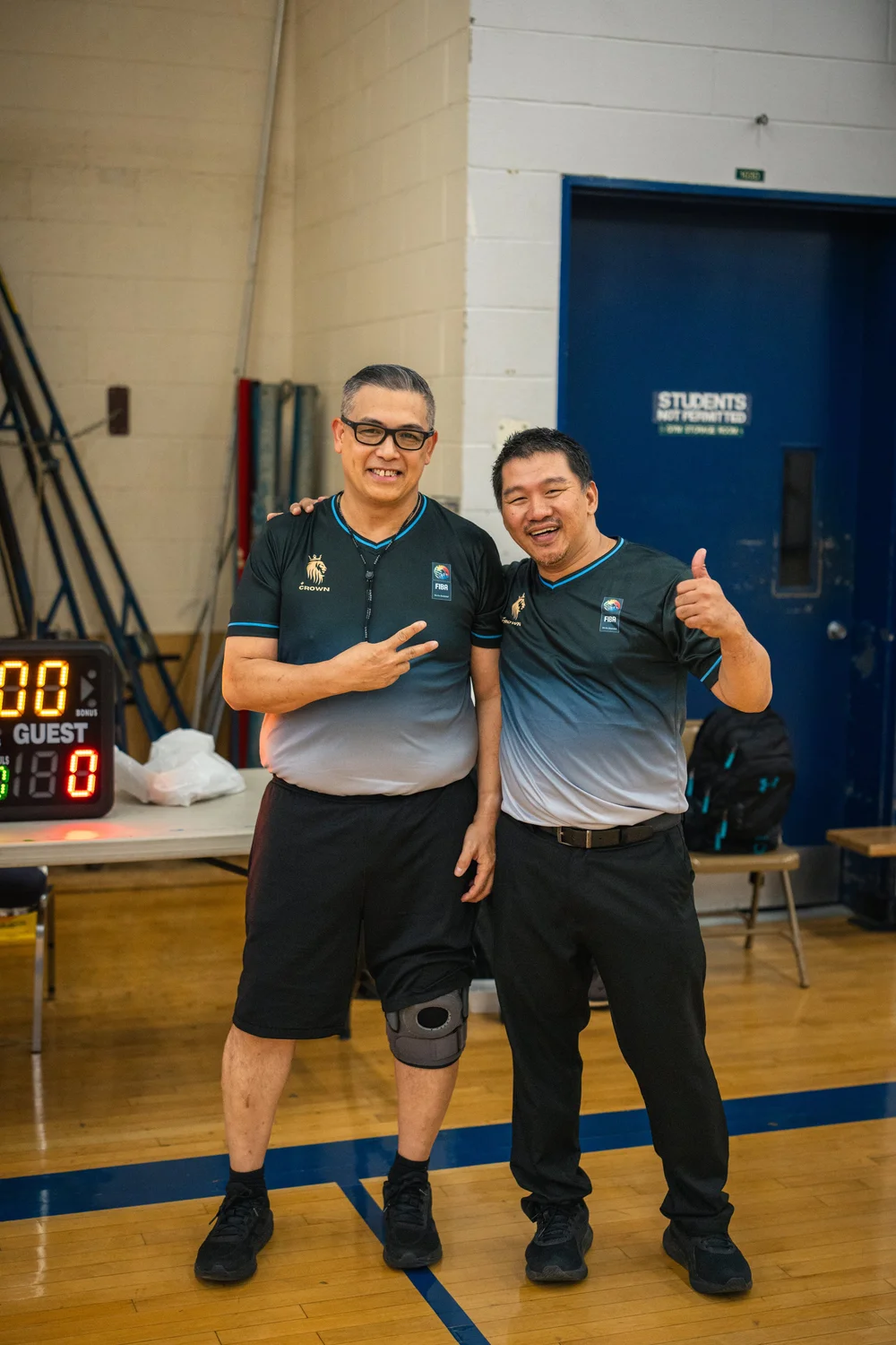 Two basketball officials pose arm in arm, one giving a peace sign and the other a thumbs-up.