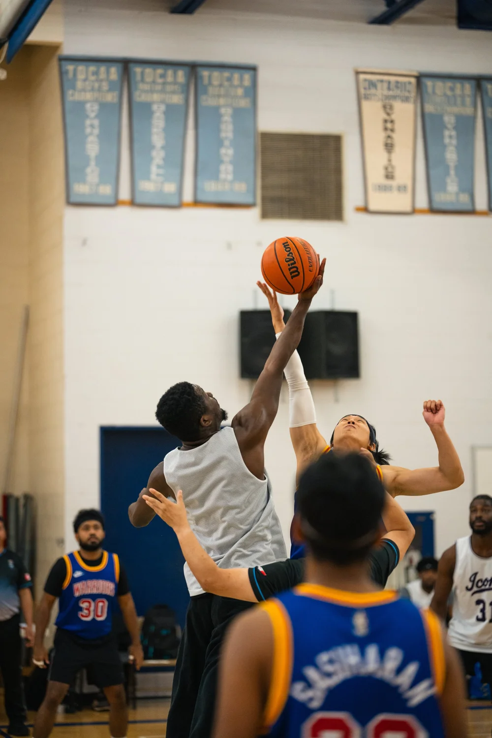 A white-shirted player and a blue-shirted player reach upward for a basketball.