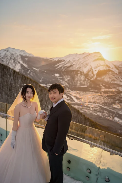 Couple at a glass railing above snowy mountains in golden light