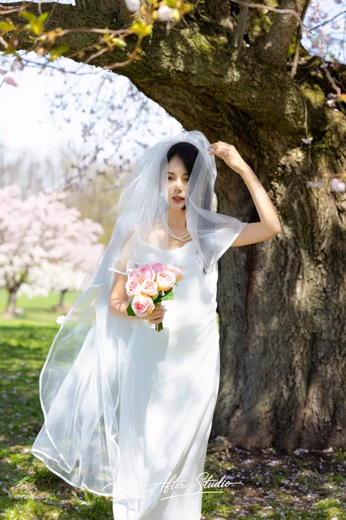 Woman with a veil and pink bouquet standing beside a broad tree trunk