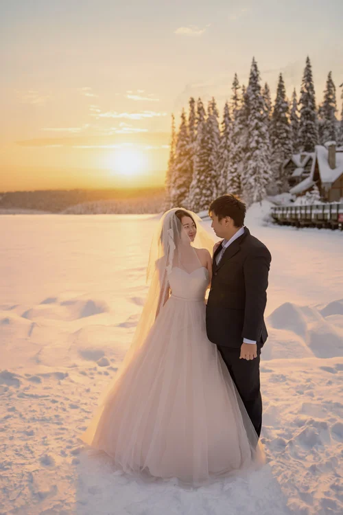 Couple in wedding attire standing beside snowy evergreens at sunset