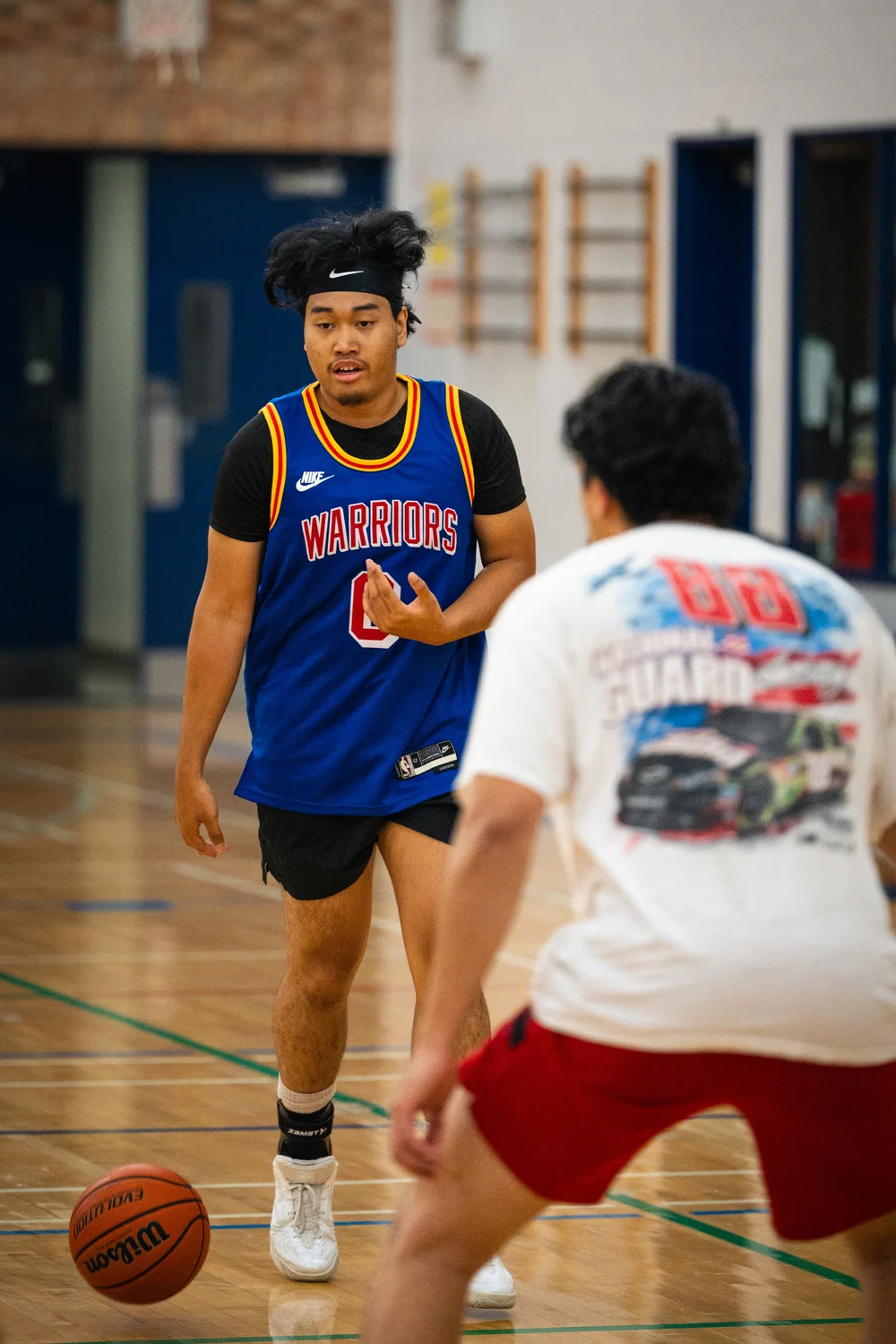Warriors number 0 dribbles toward a defender in an off-white shirt and red shorts.