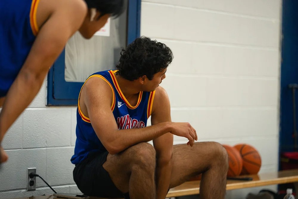 A seated Warriors player looks toward the court, with two basketballs beside him.