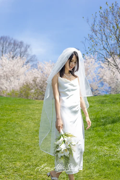 Woman holding a white bouquet on a green slope beneath flowering trees
