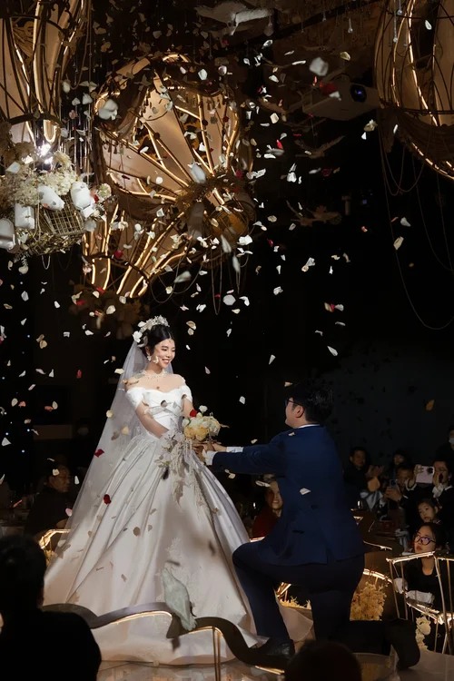 Man kneeling beside a woman in a white gown as petals fall indoors