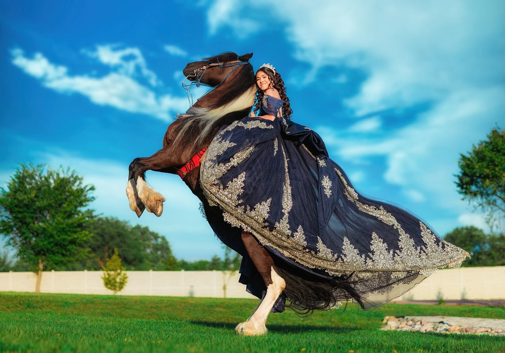 Quinceañera in a navy and gold gown posing beside a rearing horse under a blue sky