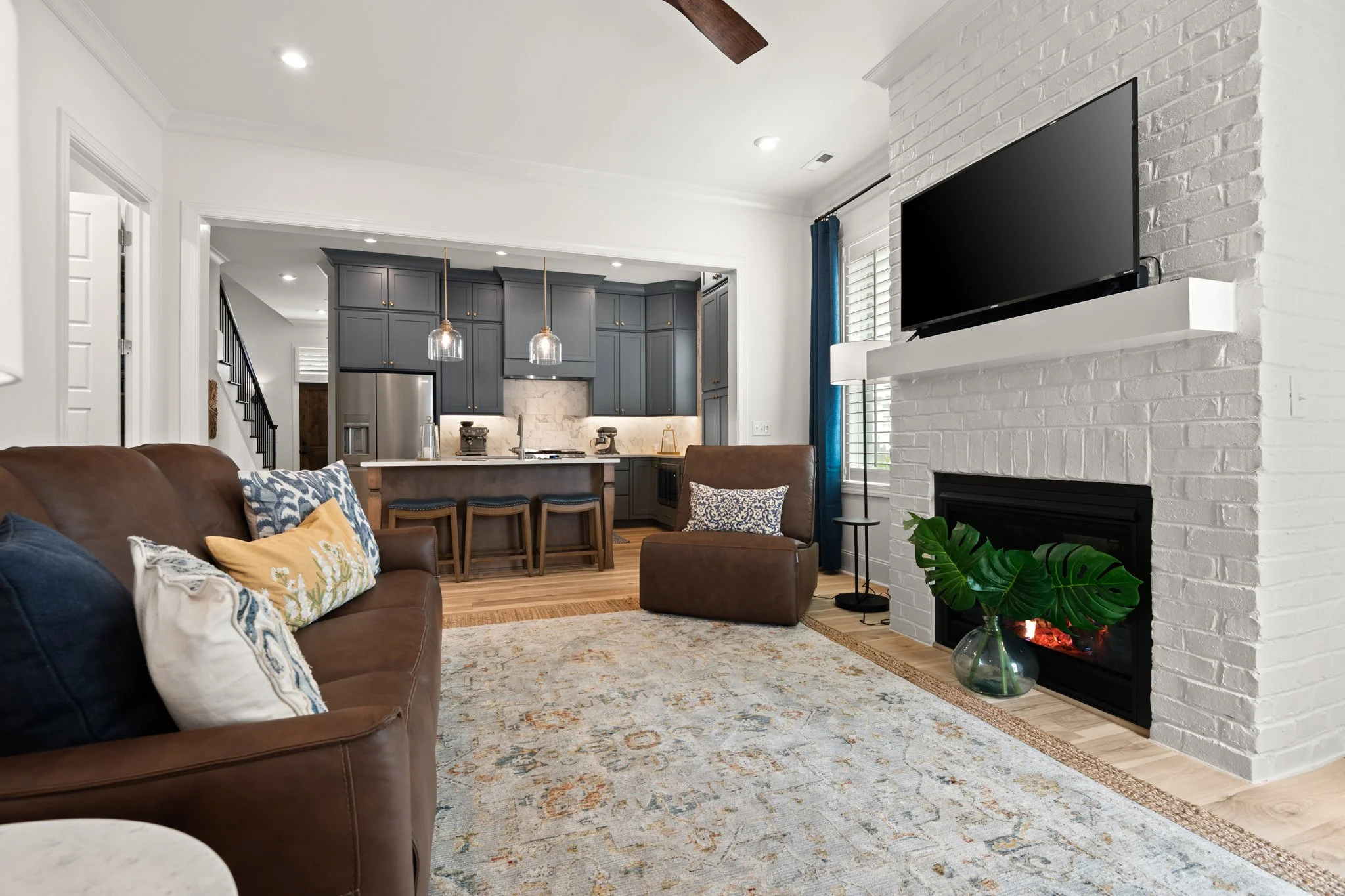 Open floor plan living room with brown leather sofa, plantation shutters, brown leather chair, fireplace with TV on mantle, patterned rug, and modern kitchen in background