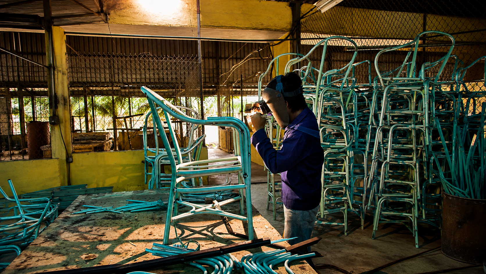 Skilled metalworker welding an aluminum furniture frame in a Vietnamese workshop with completed frames organized on racks behind