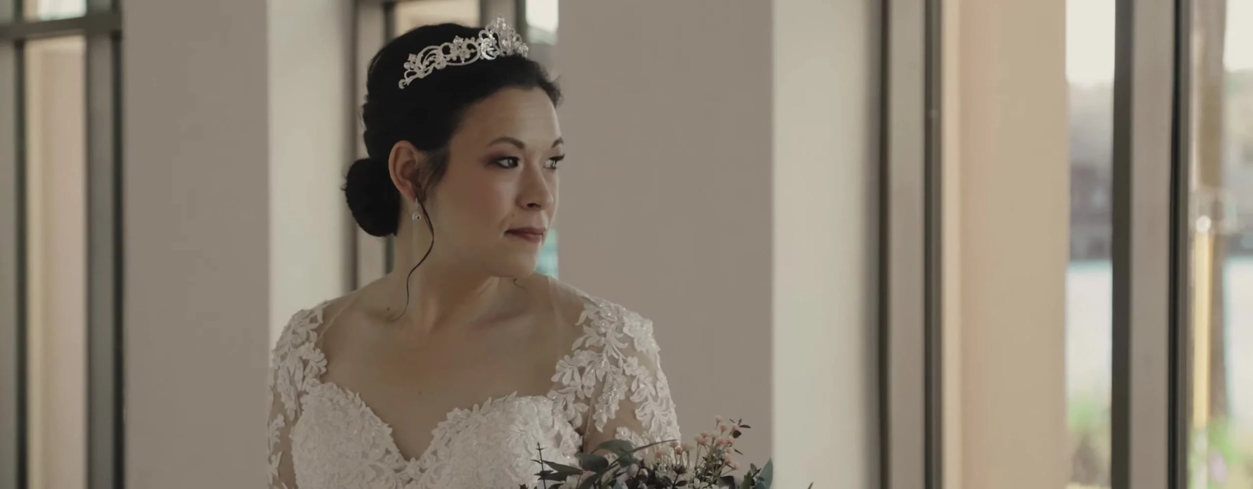 Tight portrait of the bride in a lace gown and silver tiara, looking off camera in window light