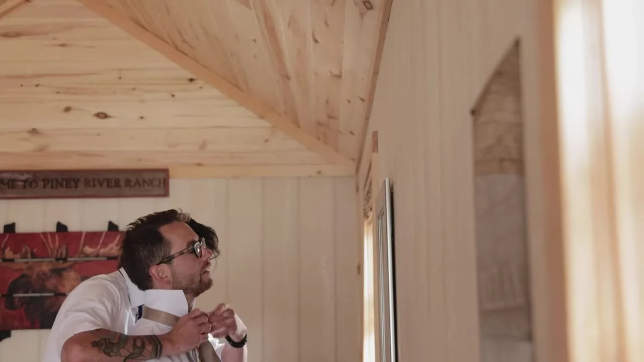 Getting ready in a Piney River Ranch cabin, hands on a champagne tie, ranch sign on the wall