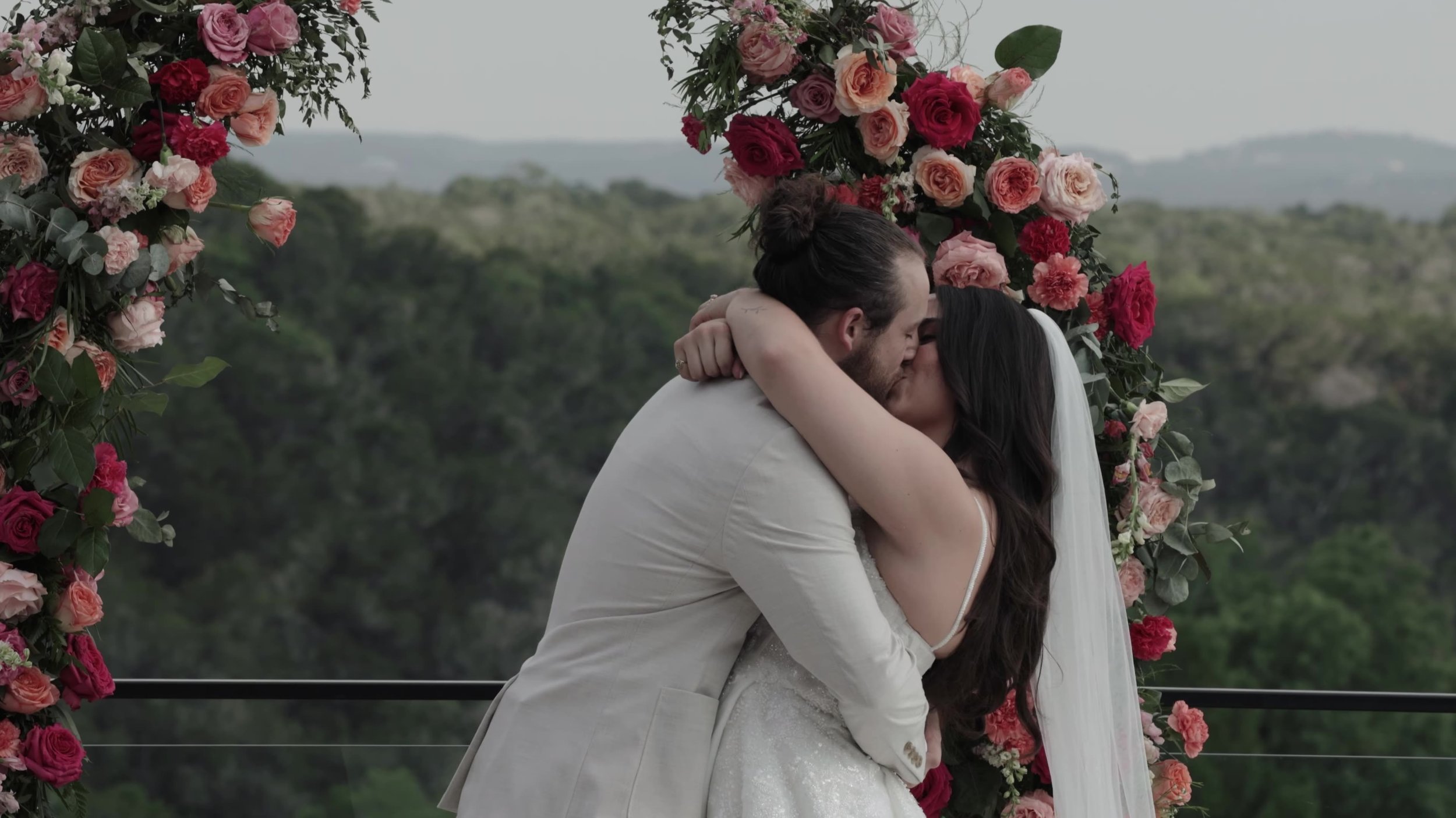 Peter and Kiyara first kiss under a pink and red floral arch at Videre Estate overlooking Austin Hill Country