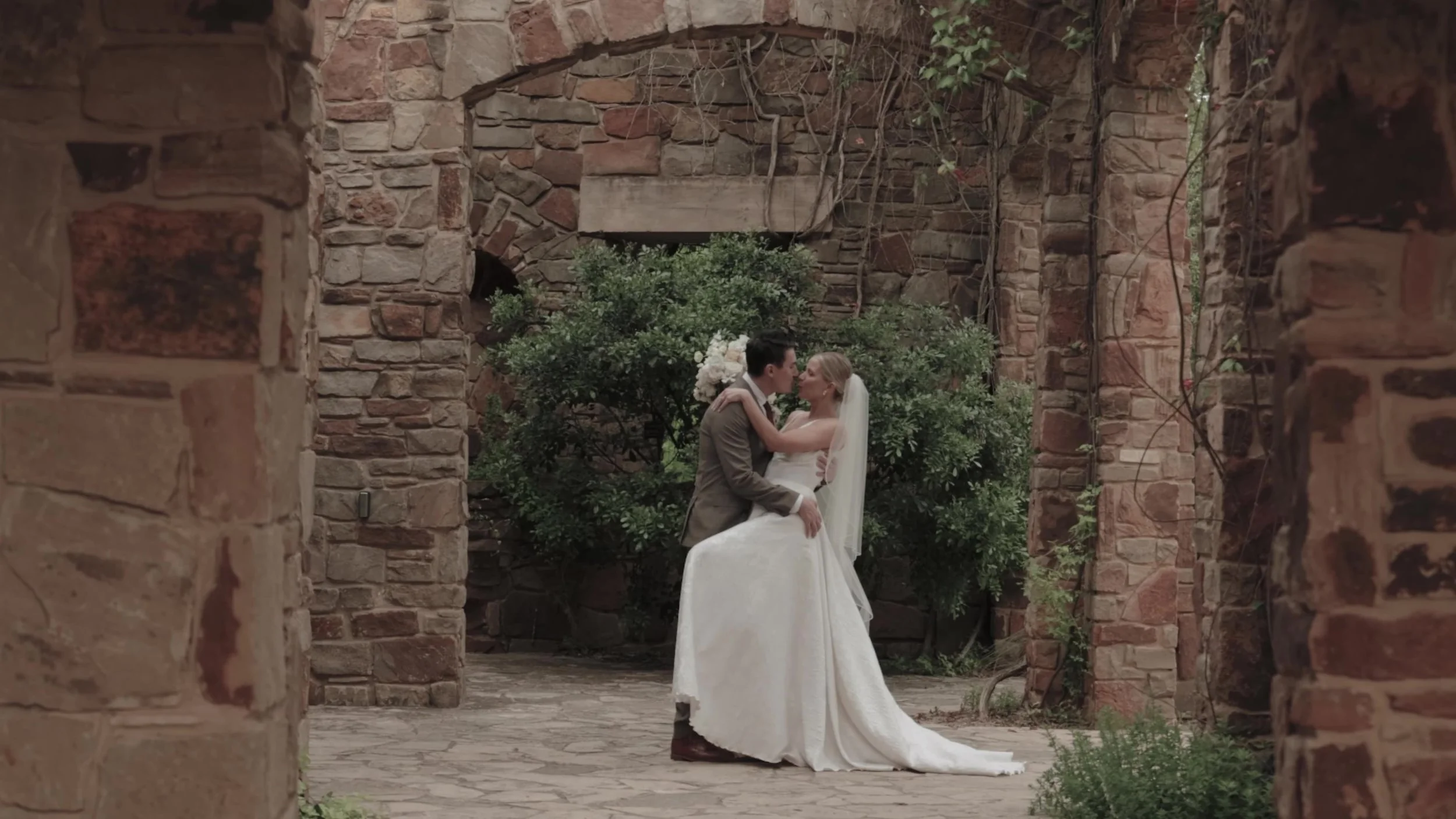 Heather and Chris kissing under a stone archway at Lady Bird Johnson Wildflower Center in Austin, Texas