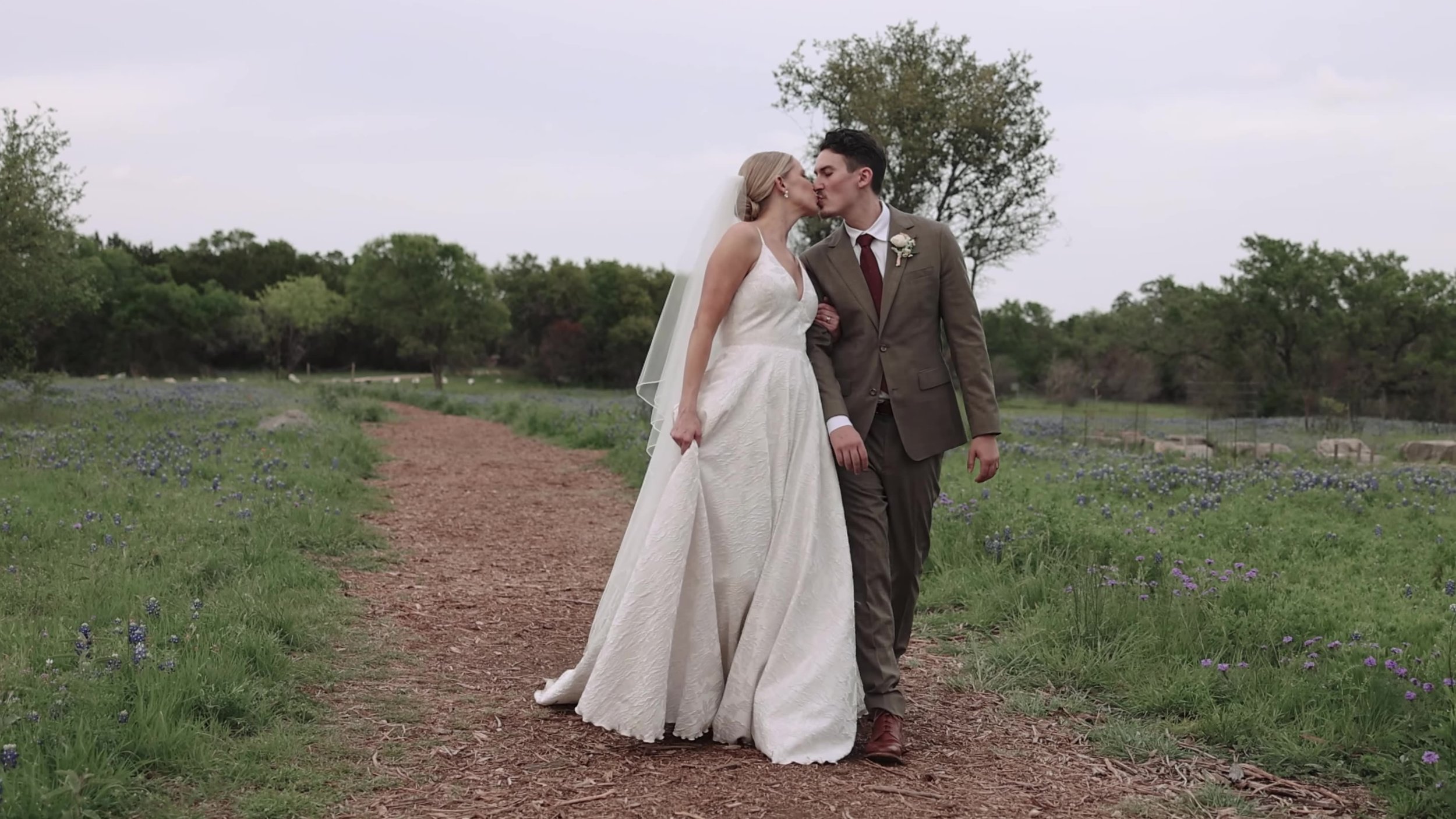 Heather and Chris walking and kissing on a path through bluebonnets at Lady Bird Johnson Wildflower Center in Austin, Texas