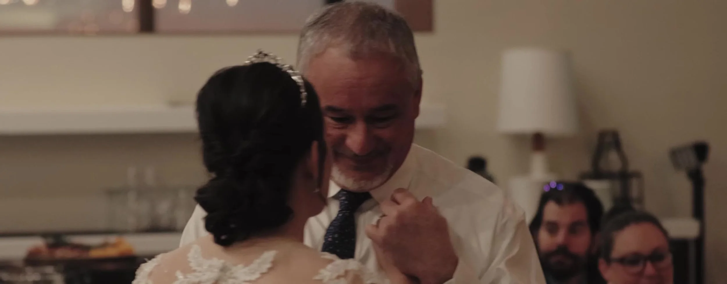 Bride dancing with her father, his face turned toward her, lace straps and a tiara in her hair