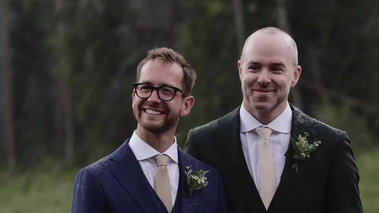Patrick and Jake standing together in navy and forest green suits, rain on their jackets, both smiling