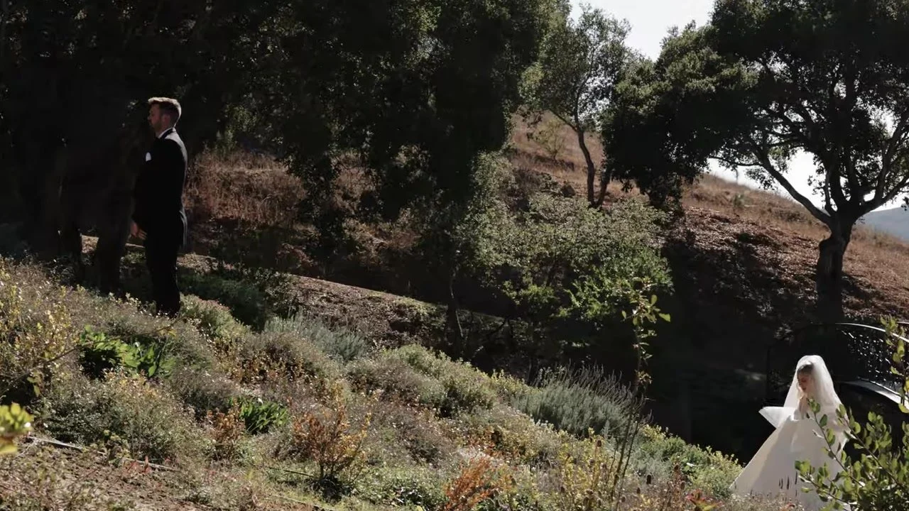 A groom in black waits under an oak while the bride in a long veil walks up a dry hillside