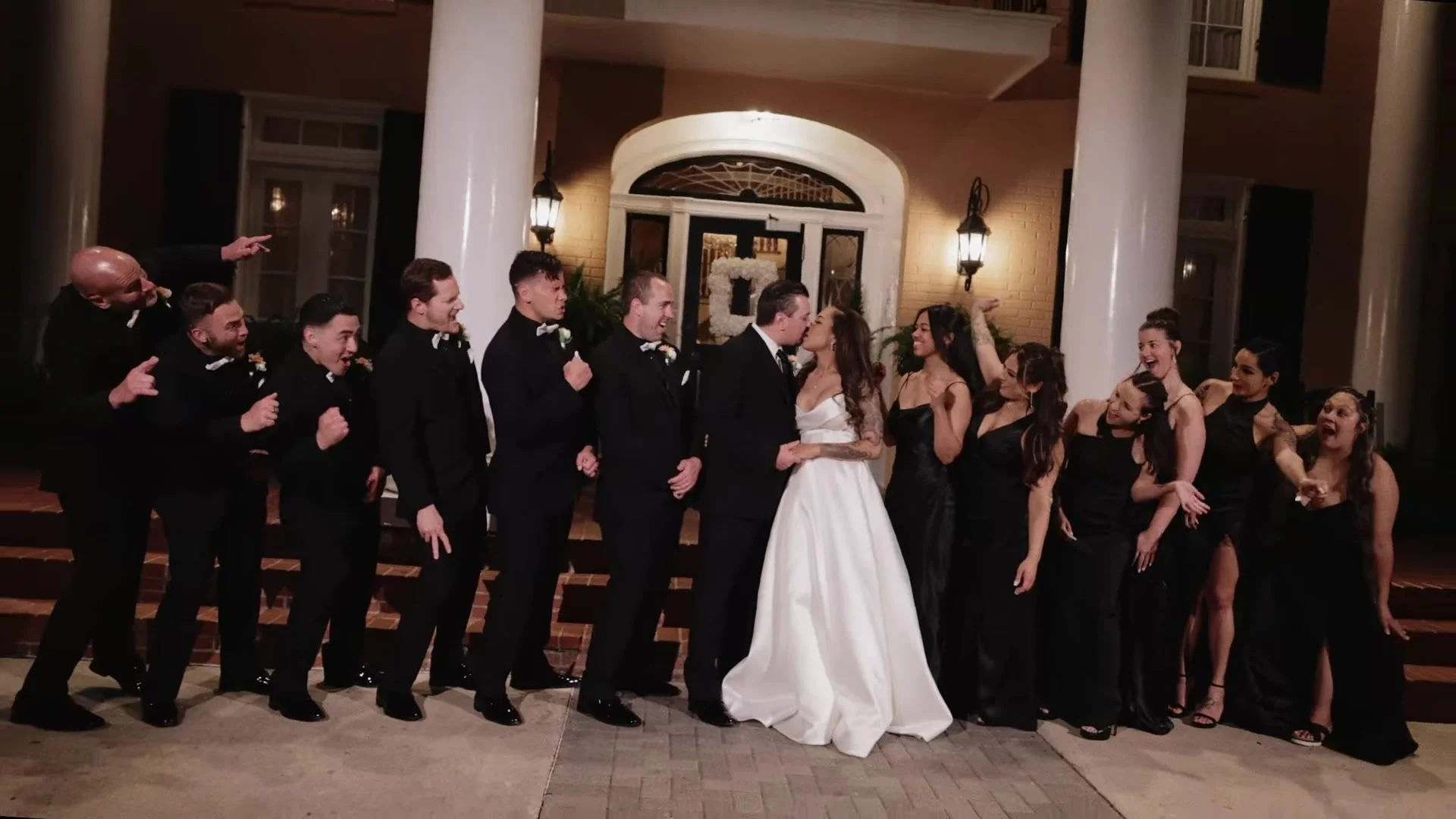 Blair and Gustavo kissing at Southern Oaks Plantation in New Orleans with wedding party cheering between white columns at night