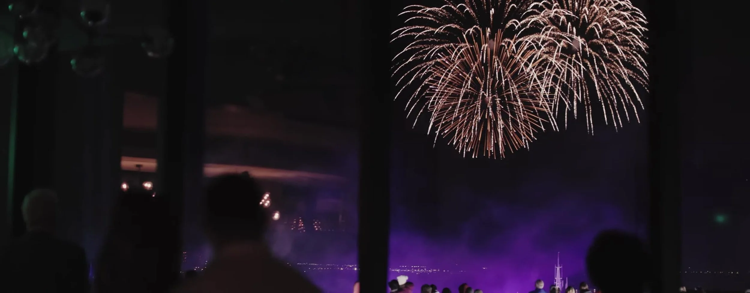 Gold fireworks over water seen through reception windows, guest silhouettes in the dark interior