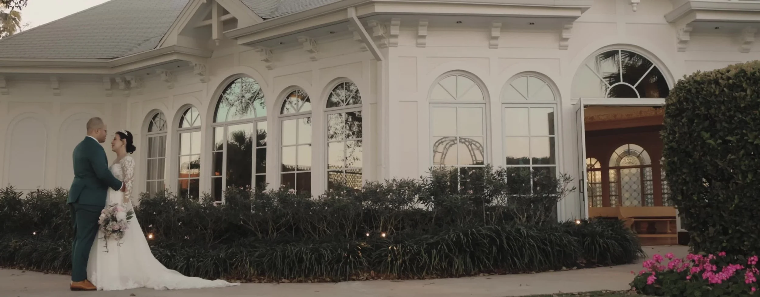 Bride and groom facing each other outside Disney's Wedding Pavilion at dusk, white arched windows and palm hedges around them