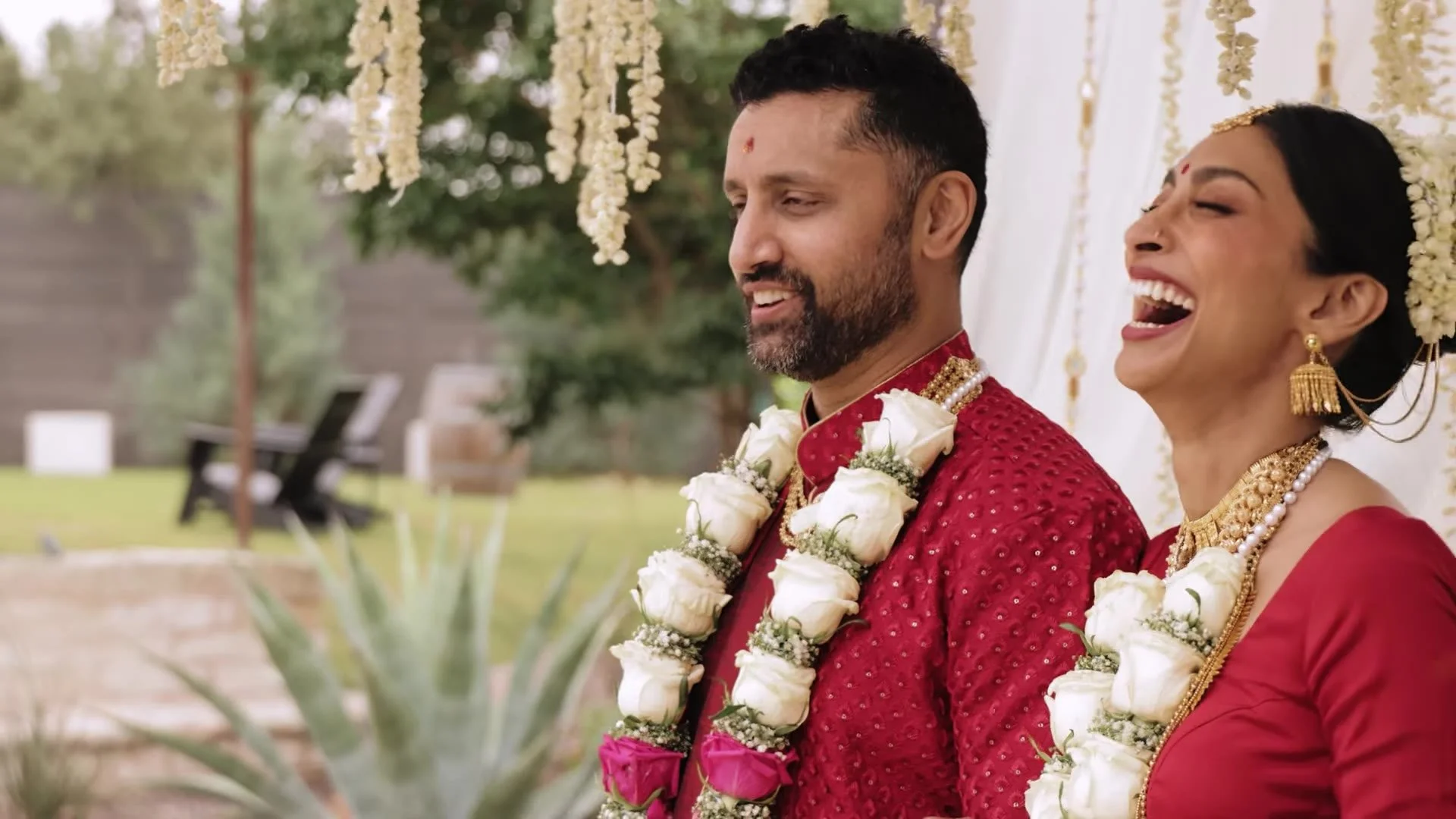 Bhavim and Mala laughing together outdoors in red Indian wedding attire with white and pink rose varmala at The Terrace Club in Austin