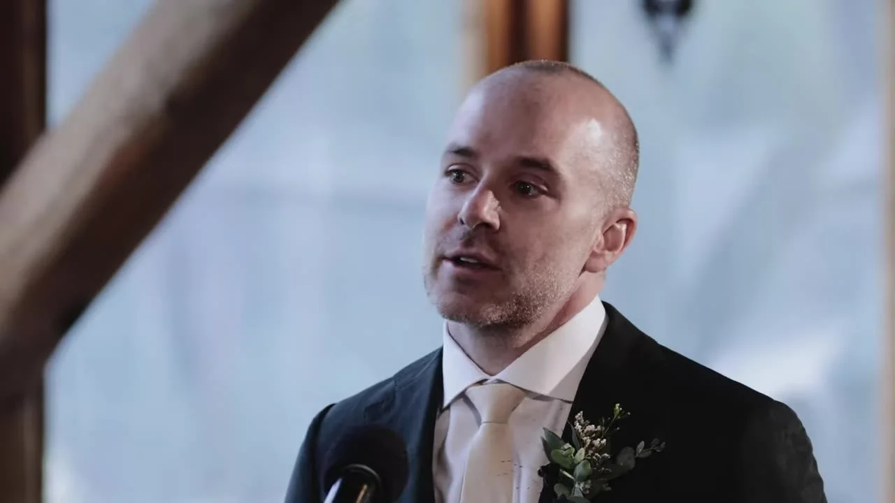 Tight portrait of a groom in a dark suit and cream tie speaking in the Piney River Ranch lodge