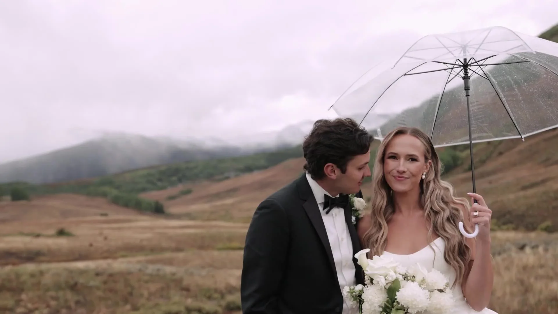 Becca and Colton under a clear umbrella at Crested Butte Wedding Garden, white bouquet and misty Colorado hills