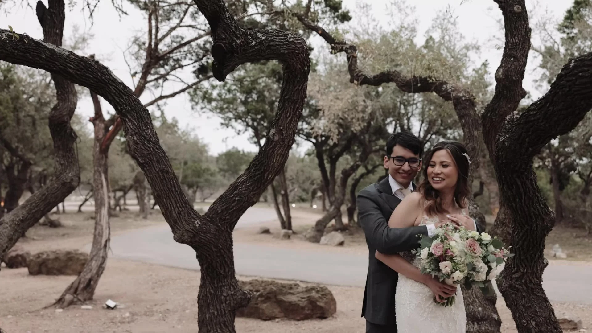 Manny hugging Jasmine from behind between oak trunks at The Terrace Club, pink and white bouquet in her hands