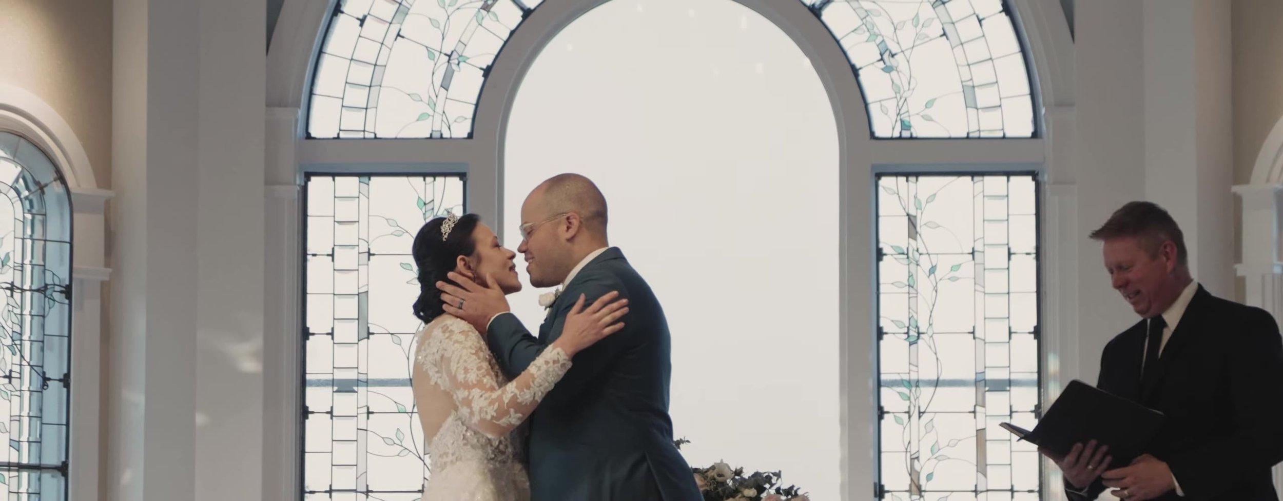 Bride and groom at the altar of Disney's Wedding Pavilion, first kiss in front of a bright arched window, officiant at right