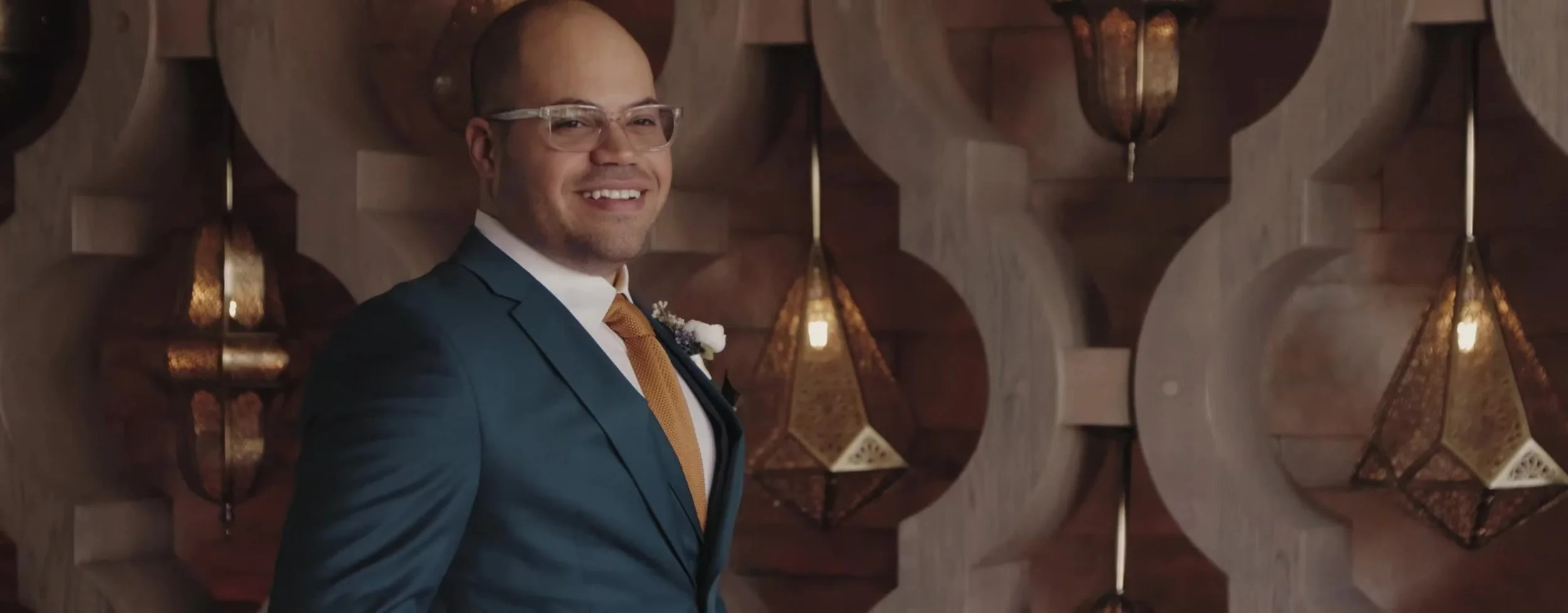 Groom in a teal suit and gold tie smiling in front of geometric wall cutouts and hanging lanterns
