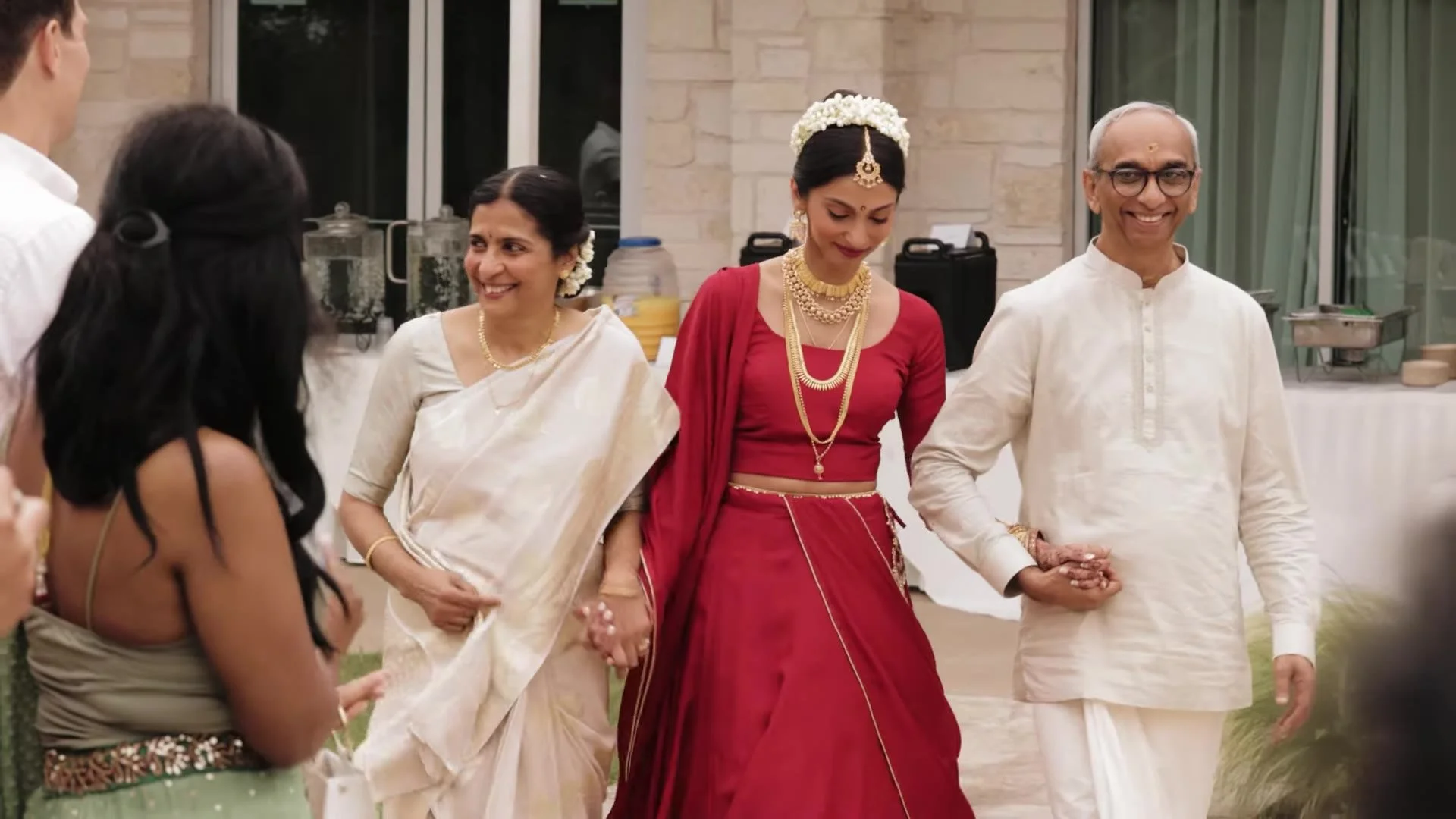 Mala in a red lehenga with gold jewelry and white jasmine gajra walking with her parents at The Terrace Club in Austin, Texas
