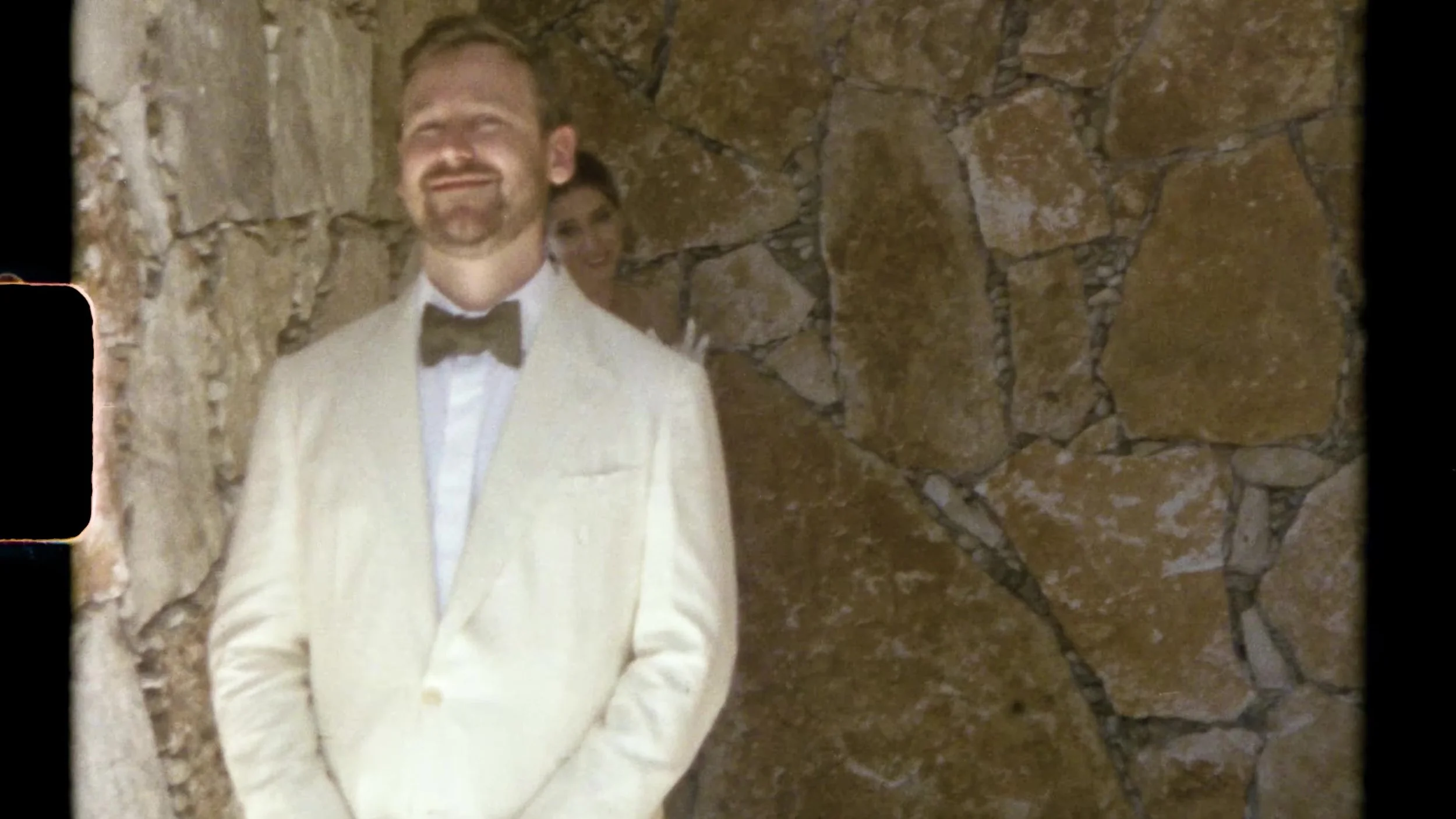 Sarah's partner in a white tuxedo and black bow tie smiling against a stone wall at Camp Lucy Vineyards