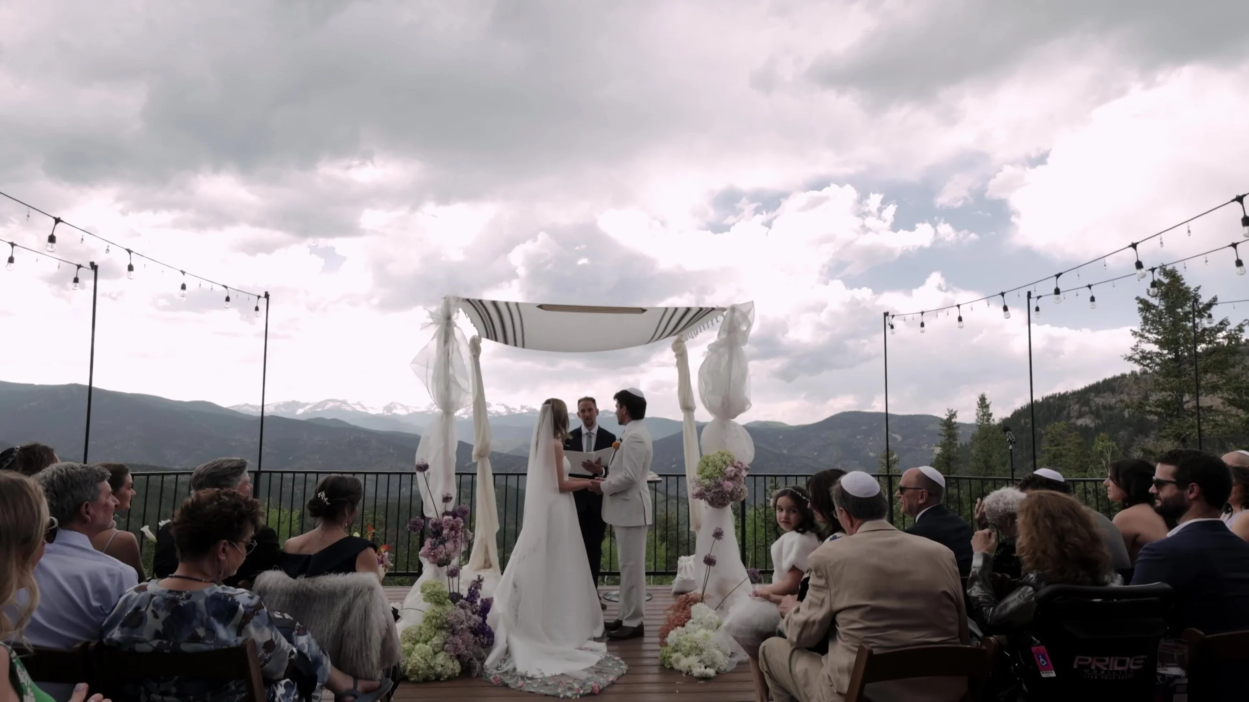 Wide outdoor ceremony under a white chuppah on the deck at Northstar Gatherings in Idaho Springs, Colorado, mountains behind the guests