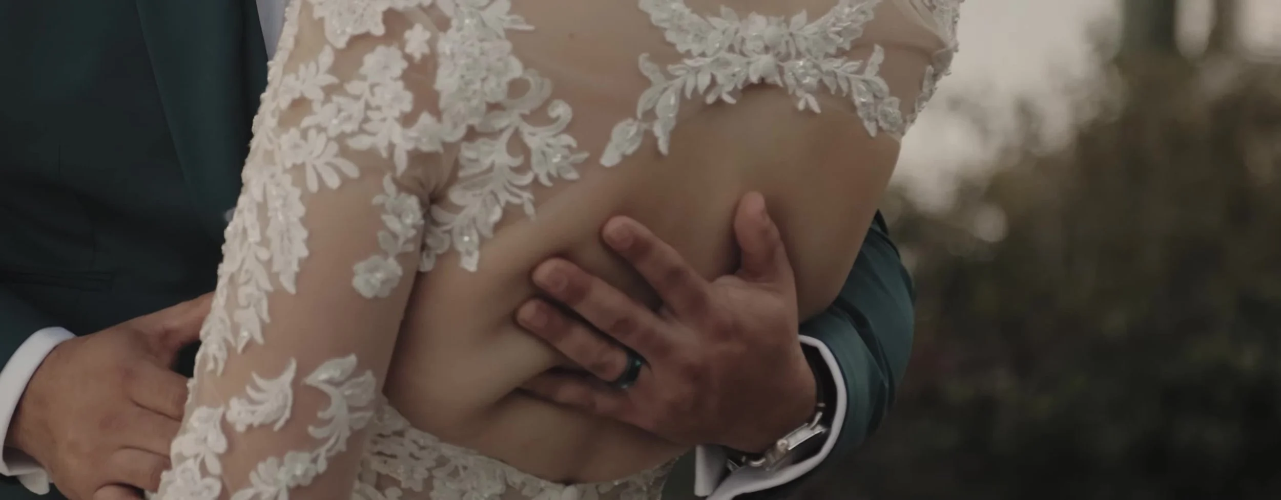 Groom's hand on the bride's lace back, dark wedding band and a watch at her waist