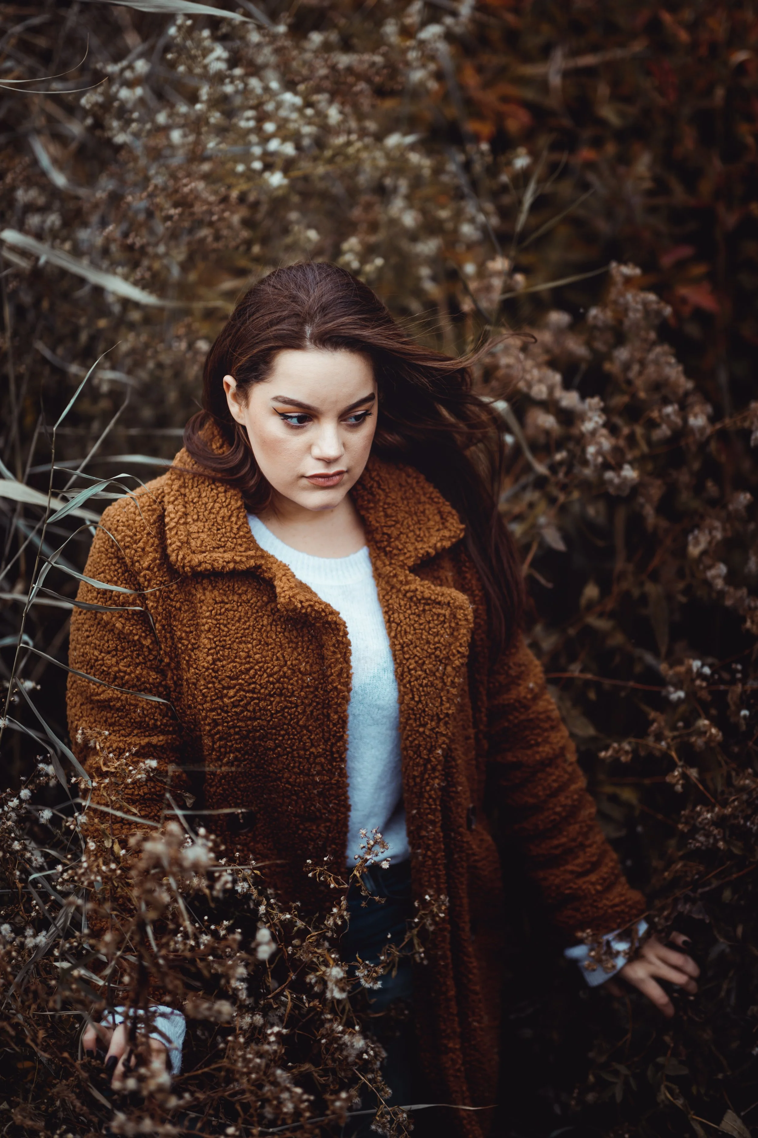Cinematic fall portrait in a warm coat surrounded by tall plants.