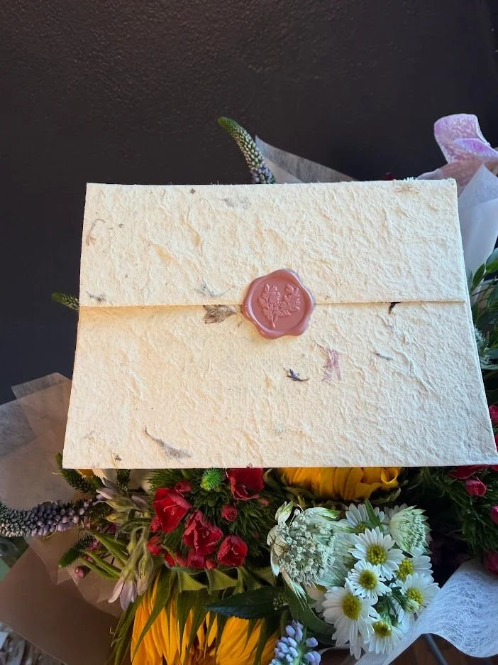 A textured paper envelope finished with a pink botanical wax seal, tucked into a bouquet.