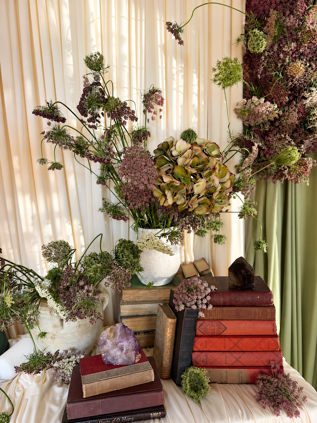 Burgundy Queen Anne’s lace and olive hydrangeas in ceramic vessels, styled with vintage books and crystals against cream draping at Esotarium.