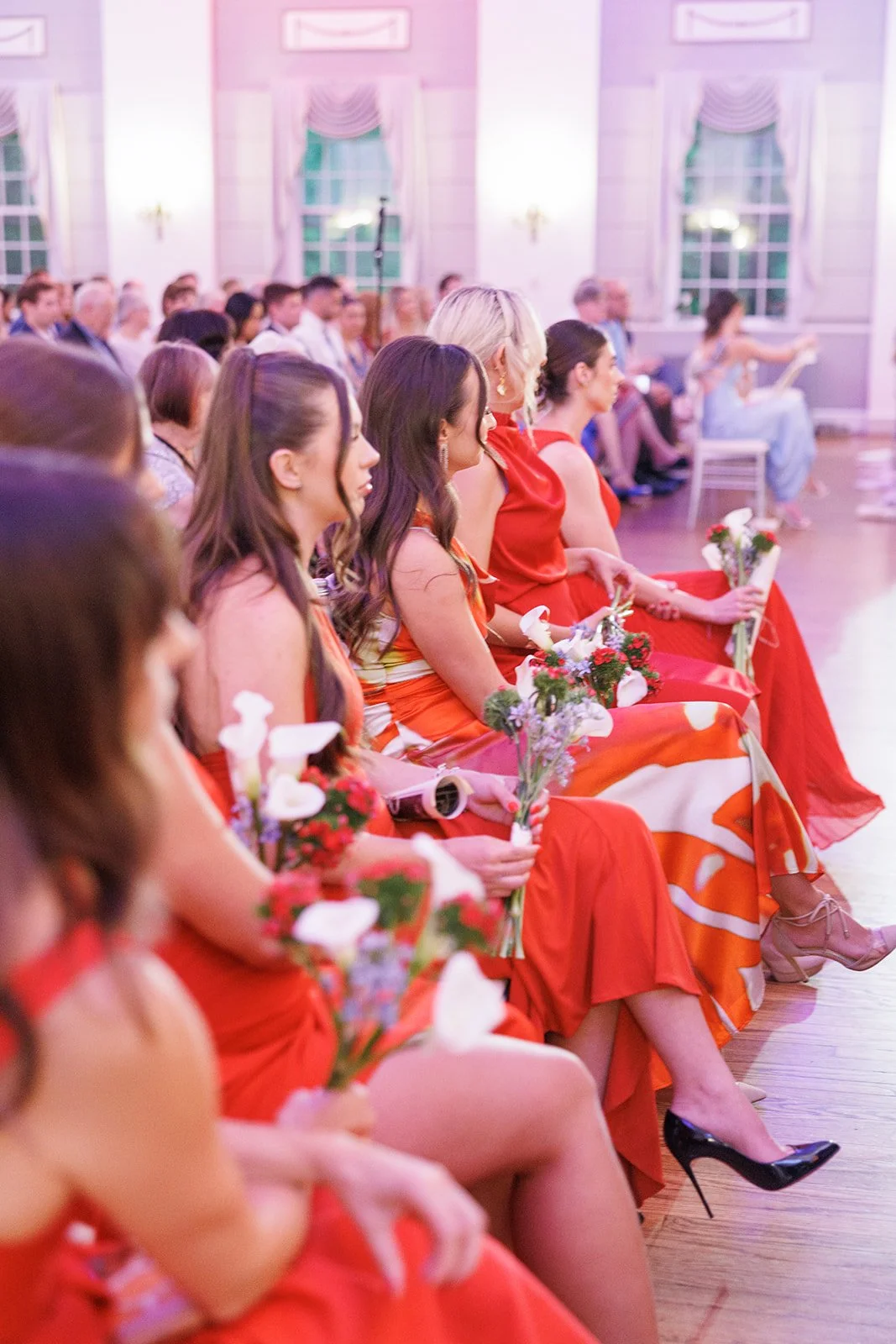Wedding party members holding small bouquets, seated in red and orange dresses.