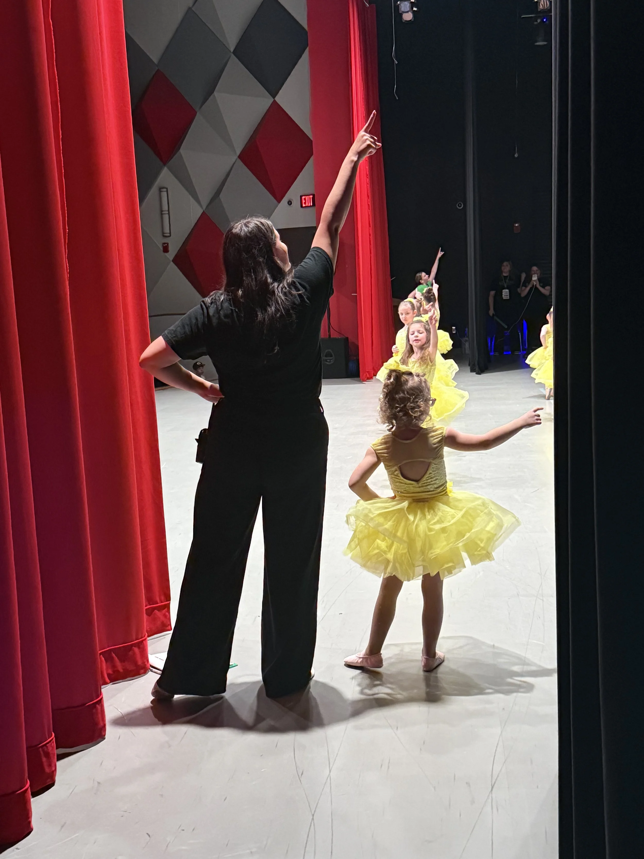 Coach Maddie guiding a little dancer in a yellow costume beside the red stage curtain