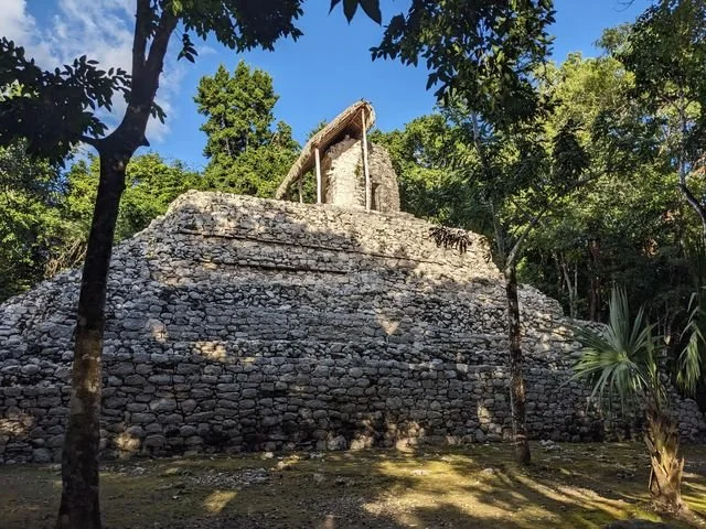 A small temple stands on a stepped stone platform among the trees at Cobá.