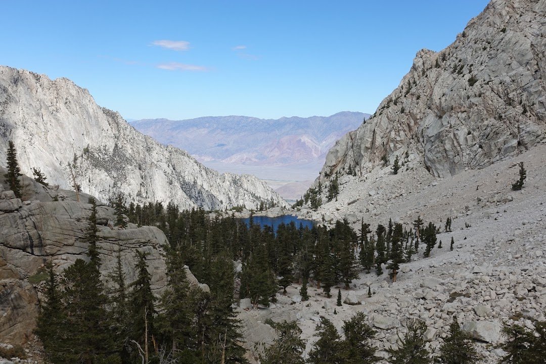 A small blue lake nestles among conifers between steep granite slopes, with the broad valley visible beyond.