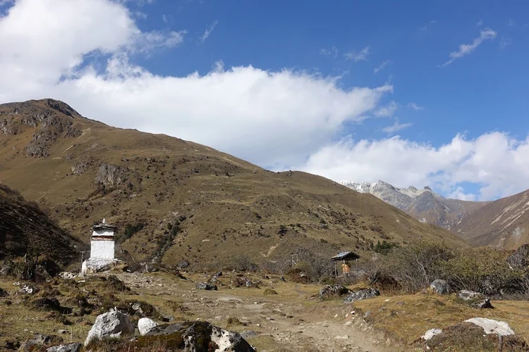 The path passes a white chorten and a small prayer-wheel shelter beneath open, brown slopes.