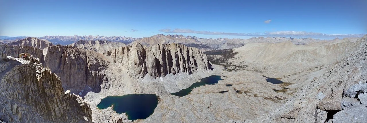 A broad panorama takes in deep blue lakes, steep cliffs and pale slopes stretching towards the distant Sierra Nevada.
