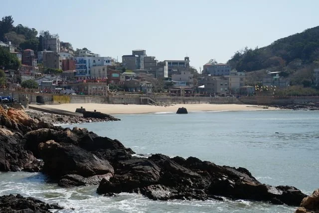 Temples and houses line a sandy bay on Nangan, above a shore of rounded rocks.