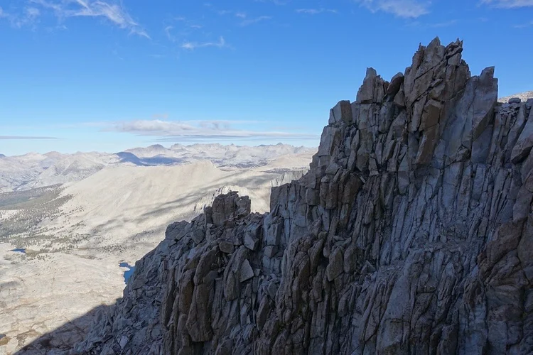 A jagged granite tower rises beside the shaded summit route, overlooking distant ridges and small alpine lakes.