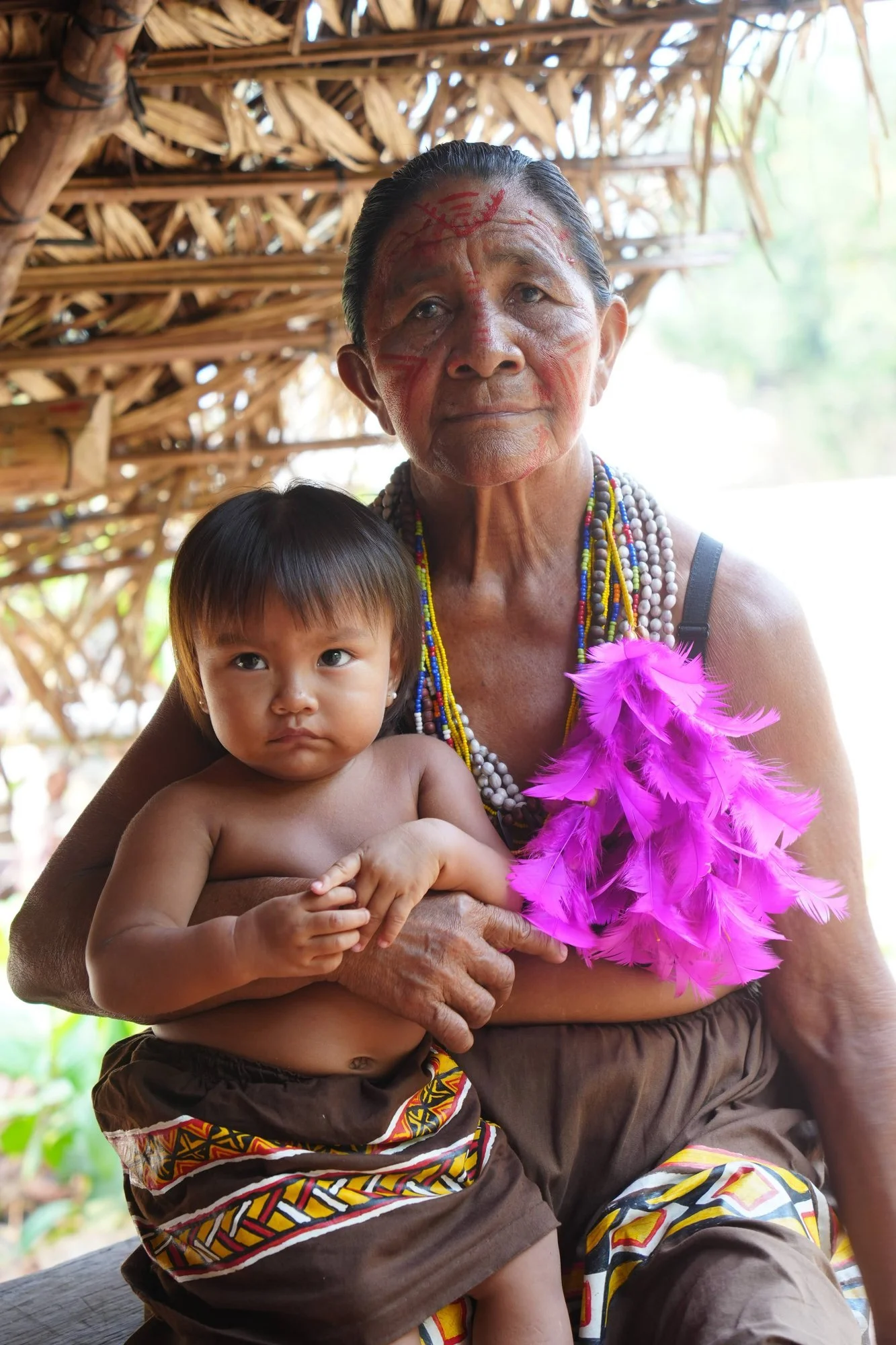 A Tuyuka woman holds a child beneath a thatched roof beside the Rio Negro, wearing bead necklaces, red face paint and vivid pink feathers.