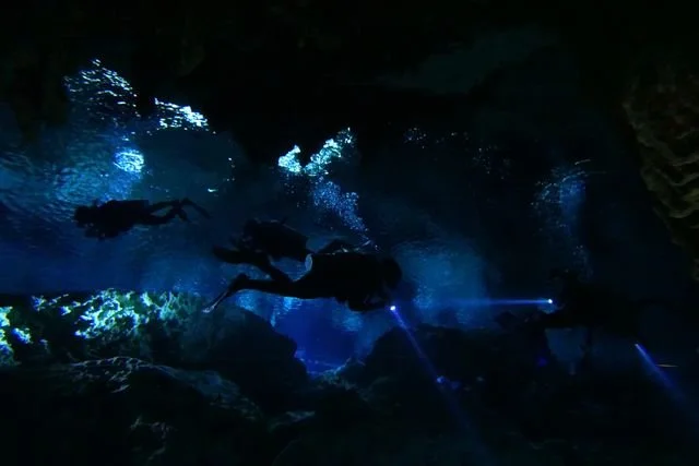 Divers cross the cavern in silhouette, their lights cutting through the blue water.