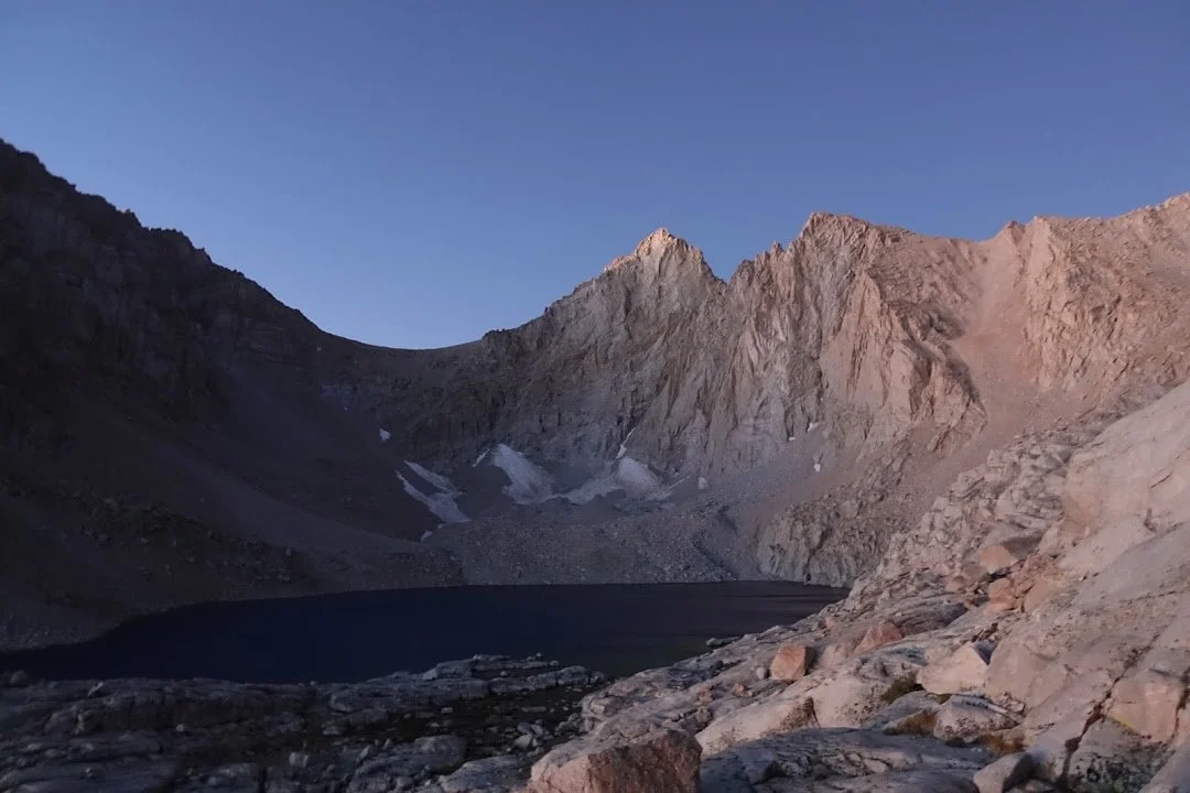 A dark alpine lake rests below steep granite cliffs, with lingering snow in the shaded gullies.