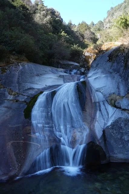 A waterfall spills over smooth pale rock into a shaded pool in the mountains near Dali.
