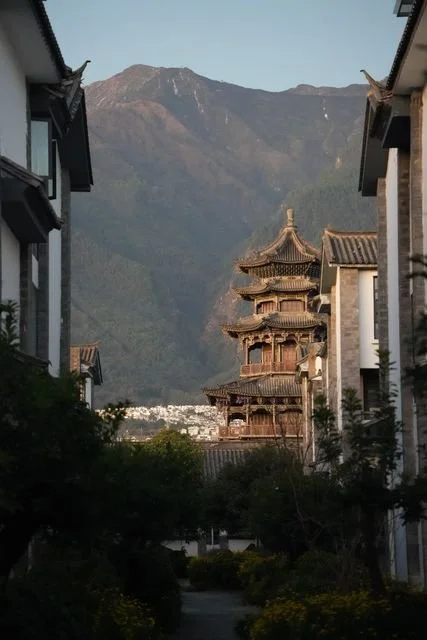 A tiered wooden building appears between white walls, beneath the mountains above Dali.